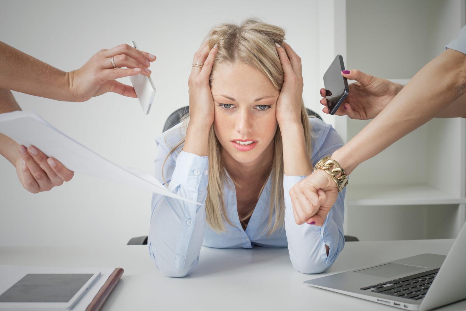 Stressed woman at desk surrounded by multiple demands, holding her head with hands